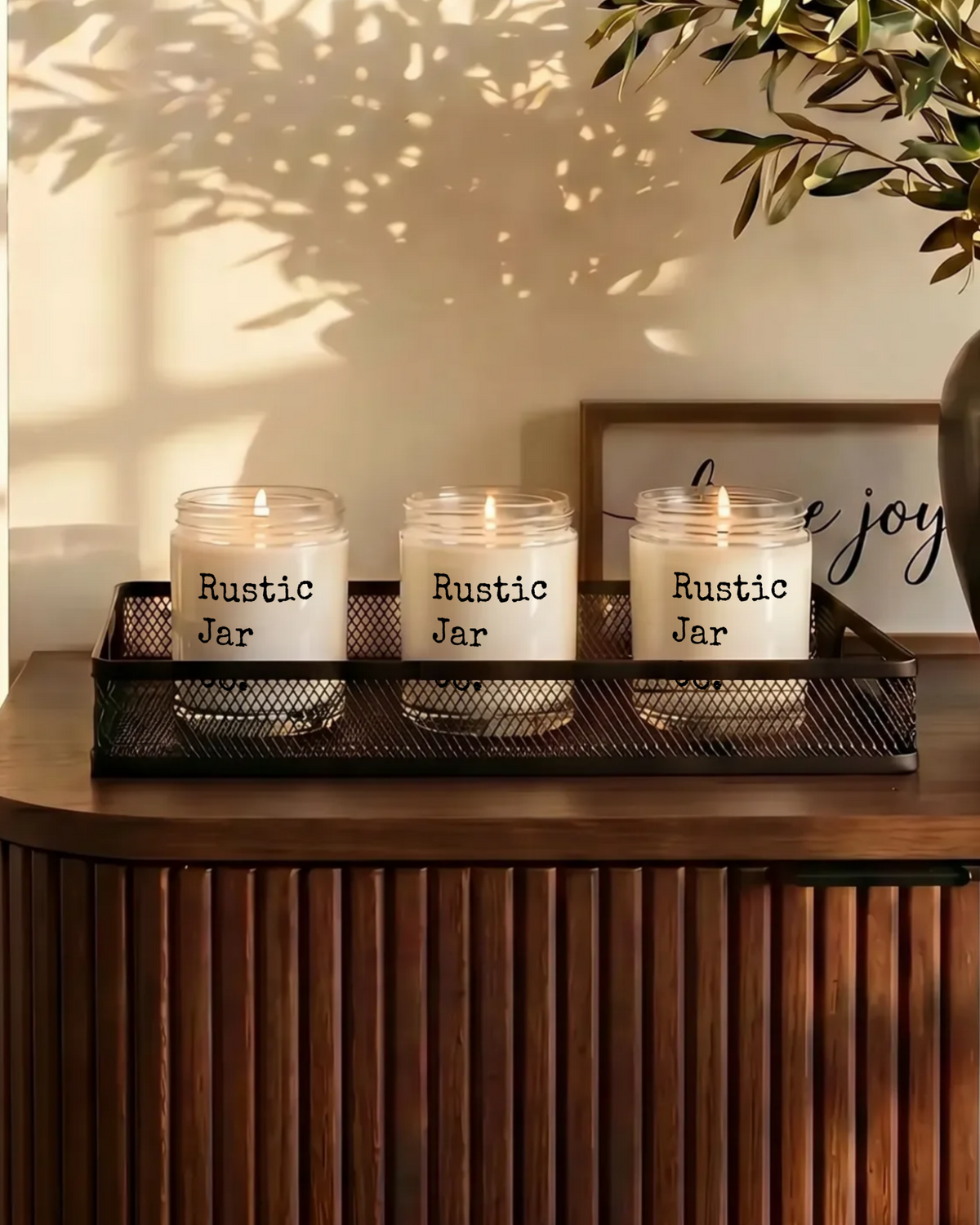 Three lit candles in clear glass jars labeled 'Rustic Jar' sit on a black metal tray on a wooden console table in a warm, sunlit room. A person is partially visible on the left, and a plant and framed sign decorate the background.